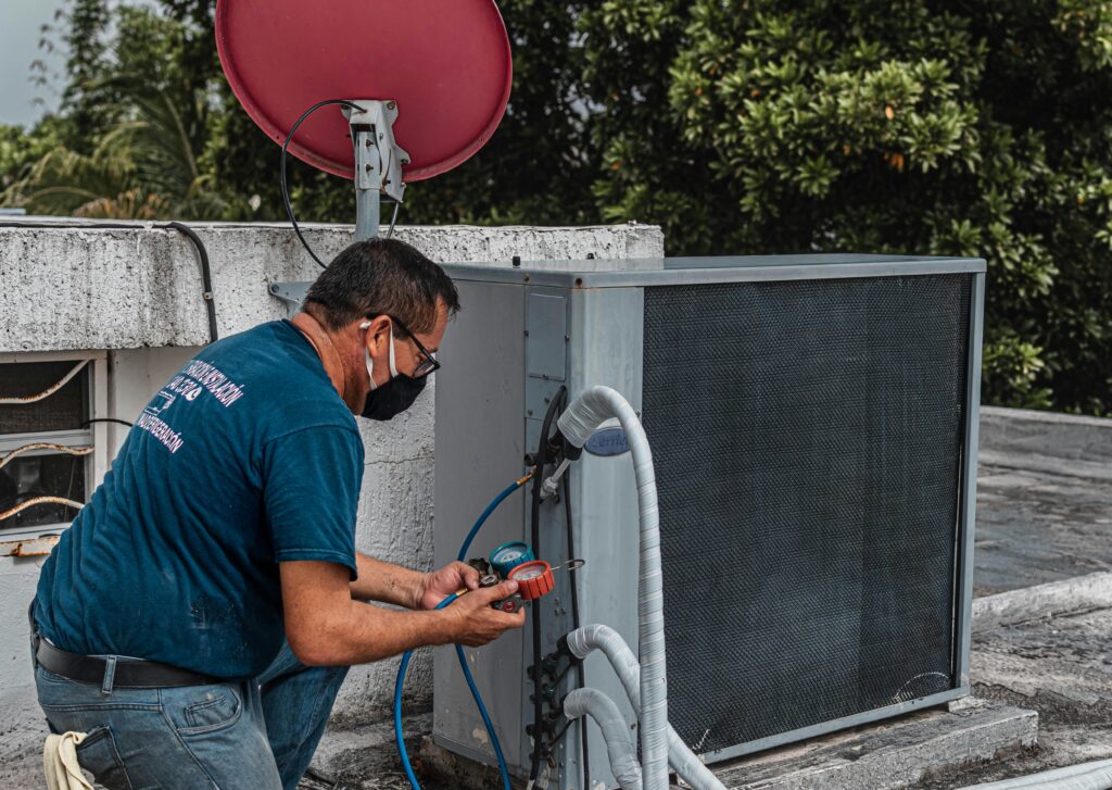 A professional HVAC technician wearing a blue shirt and a face mask is servicing an outdoor air conditioning unit on a rooftop. The technician is using tools and gauges connected to the unit, with coiled insulated pipes and a satellite dish visible in the background. The setting includes lush greenery in the distance.HVAC maintenance tips